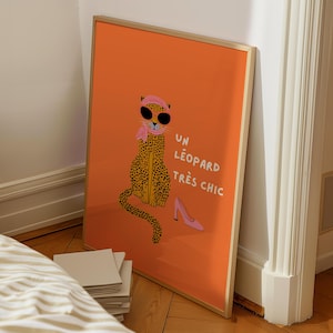 Op de afbeelding: Ingelijste kunstprint met een oranje achtergrond, met een cartoon luipaard met zonnebril, een roze sjaal en een hoofdband. Er staat een roze high heel schoen naast het luipaard. De Franse tekst "UN L&Eacute;OPARD TR&Egrave;S CHIC" staat ook op de print.
