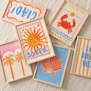 Op de afbeelding: Een verzameling ingelijste kunstprints met een zomerthema. De prints bevatten illustraties van een krab, een zeeschelp, palmbomen, een lachende zon en zinnen als "Ciao" en "La Dolce Vita". Het kleurenpalet omvat oranje, blauw, roze en geel.
