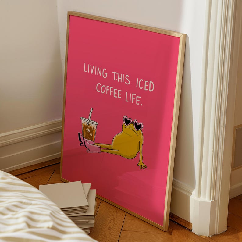 Op de afbeelding: Een ingelijste roze kunstprint met de tekst "LIVING THIS ICED COFFEE LIFE." De illustratie toont een gele kikker met hartvormige zonnebril en roze laarzen, die een ijskoffie vasthoudt. De lijst is goudkleurig.