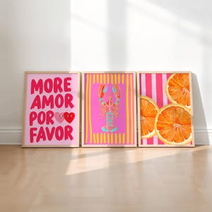 May include: Three framed art prints with a light wood finish. The prints feature a pink background with the words "MORE AMOR POR FAVOR" and two hearts, a pink lobster, and orange slices. The prints are displayed on a light wood floor.