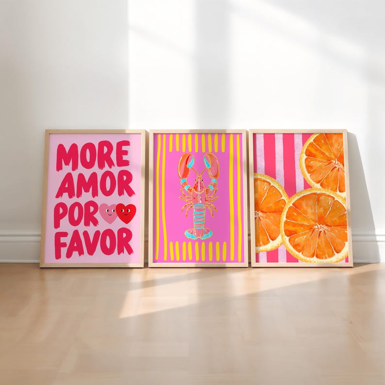 May include: Three framed art prints with a light wood finish. The prints feature a pink background with the words "MORE AMOR POR FAVOR" and two hearts, a pink lobster, and orange slices. The prints are displayed on a light wood floor.