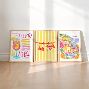 May include: Three framed art prints. The left print features a cocktail with the words "SPRITZ APEROL". The center print shows a bikini hanging on a clothesline against a yellow striped background. The right print depicts a coastal town with the word "Amalfi".