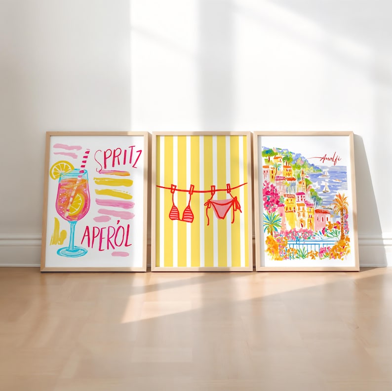 May include: Three framed art prints. The left print features a cocktail with the words "SPRITZ APEROL". The center print shows a bikini hanging on a clothesline against a yellow striped background. The right print depicts a coastal town with the word "Amalfi".