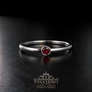 May include: A silver ring with a round, deep red gemstone. The ring has a thin band and a simple, elegant design. The gemstone is set in a bezel setting. The ring is displayed on a dark surface.