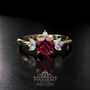 May include: A gold ring featuring a large, round, deep red gemstone as the centerpiece, accented by smaller white gemstones. The ring has a delicate, floral-inspired design with the brand name "Pissarro Fine Jewelry" at the bottom.