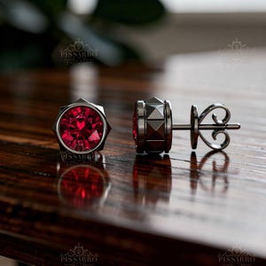 May include: A pair of stud earrings featuring a round, faceted, vibrant red gemstone set in a dark metal frame. The side of the earring setting displays a geometric, pyramid-like stud pattern, and the back includes a secure butterfly-style post.