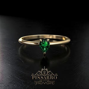 May include: A gold ring featuring a pear-cut, emerald-green gemstone. The ring is set against a dark backdrop, with the PISSARRO FINE JEWELRY logo below. The ring's band is smooth and polished.