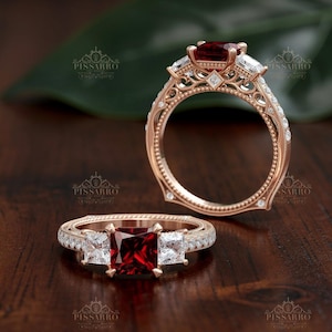 May include: Two rose gold rings with intricate filigree detailing. One ring features a large, square, deep red gemstone flanked by two smaller white gemstones. The other ring has a similar design, with a large red gemstone.