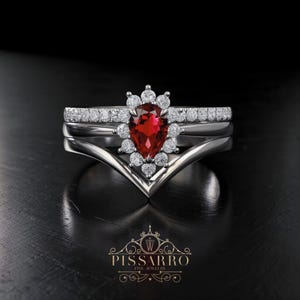 Op de afbeelding: Een driedelige zilveren ringenset. De middelste ring heeft een peervormige, dieprode edelsteen omringd door kleine, ronde witte edelstenen. De andere twee ringen zijn zilveren banden, een met kleine witte edelstenen. De ringen liggen op een zwart oppervlak.