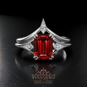 May include: A silver ring with a large, rectangular, deep red gemstone as the centerpiece. The ring features a unique design with smaller, clear gemstones and a pointed top. The brand name "Pissarro Fine Jewelry" is displayed at the bottom.