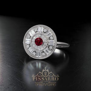 May include: A silver ring with a circular design, featuring a central red gemstone surrounded by diamonds and baguette-cut stones. The ring has a polished band and is set against a dark background. The ring is a piece of fine jewelry.