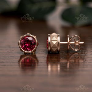Könnte beinhalten: Ein Paar Ohrstecker aus Roségold mit einem runden, facettierten, dunkelrosa Edelstein in einer Zargenfassung. Die Seite des Ohrsteckers zeigt ein texturiertes Pyramidenmuster und der Verschluss ist als sicherer Schmetterlingsverschluss ausgeführt.