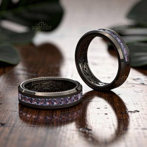 May include: Two black rings with a row of square, light purple gemstones. The rings have an intricate, patterned design on the inside band. The rings are displayed on a wooden surface.