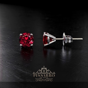 May include: A pair of stud earrings featuring round, faceted, deep red gemstones set in a silver metal. The earrings are displayed on a dark surface, with the brand logo "Pissarro Fine Jewelry" visible.