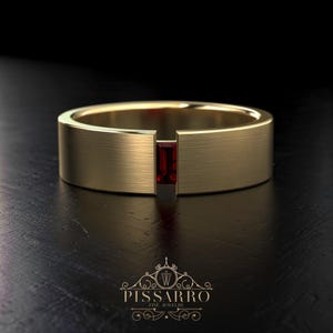 May include: A gold-colored ring with a rectangular, deep red gemstone set in the center. The ring has a brushed metal texture and a modern, minimalist design. The Pissarro Fine Jewelry logo is at the bottom.