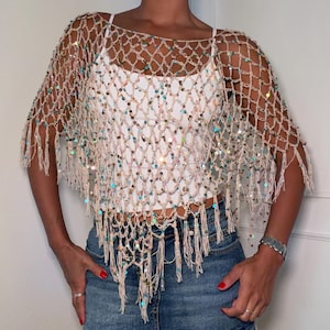 Puede incluir: Un poncho de red de ganchillo en color beige con lentejuelas iridiscentes por todo el tejido de punto abierto. La prenda tiene un escote amplio, mangas cortas y un borde con flecos, superpuesta sobre una camiseta blanca sencilla y una falda vaquera azul.