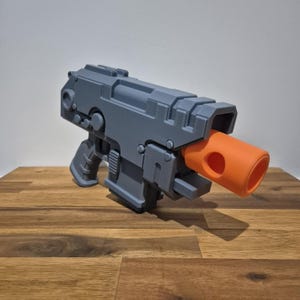 以下が含まれることがあります： オレンジ色の銃身を持つグレーの3Dプリントされたおもちゃの銃。銃は、テクスチャ加工された表面を持つ未来的なデザインです。
