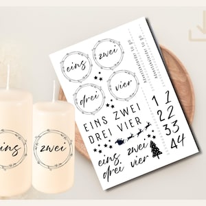 Puede incluir: Dos velas de pilar de color marfil con las palabras alemanas "eins" y "zwei" en un diseño de corona circular. Una hoja de pegatinas de calendario de Adviento presenta números, estrellas y las palabras "EINS ZWEI DREI VIER".
