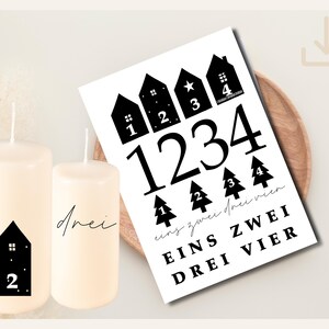 Puede incluir: Dos velas de pilar de color marfil con gráficos de casas negras y números, junto a una impresión en blanco y negro con números y palabras alemanas. La impresión presenta los números 1, 2, 3 y 4, y las palabras alemanas "Eins, Zwei, Drei, Vier."