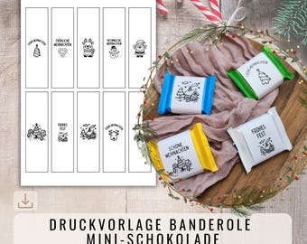 May include: A sheet of black and white Christmas-themed labels with festive illustrations and text. Beside it, wrapped mini chocolate bars in blue, green, yellow, and white, also with Christmas designs. A candy cane and greenery complete the holiday theme.