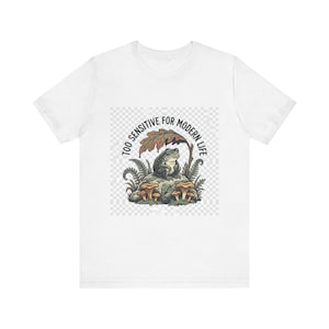 May include: A white t-shirt featuring a graphic of a green frog sitting on a rock under a large leaf, surrounded by mushrooms and ferns. The text "TOO SENSITIVE FOR MODERN LIFE" is printed in an arc above the frog, set against a light checkerboard background.