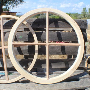 Puede incluir: Dos marcos de ventana circulares de madera sin terminar con un diseño interior en forma de cruz. Los marcos son de madera de color claro y están colocados contra un palé de madera desgastado. La escena es al aire libre, con luz natural y un fondo rural.