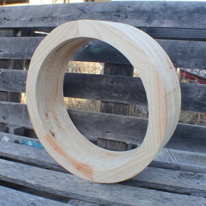May include: A circular wooden frame, likely for a wreath or decorative element, is displayed. The unfinished wood has a natural, light color with visible grain and some reddish-brown accents. The frame is resting on a weathered wooden pallet.