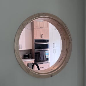 Pode incluir: Um espelho de parede circular com uma moldura grossa de madeira clara natural com padrões de veios visíveis. O espelho reflete o interior de uma cozinha, mostrando armários brancos e de madeira, um forno duplo e uma bancada com aparelhos.