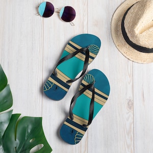 Op de afbeelding: Een paar blauwe en turquoise slippers met een schelpdesign en zwarte bandjes. De slippers liggen op een wit houten oppervlak.
