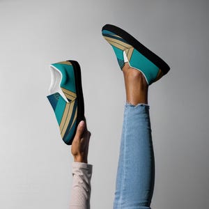 Pode incluir: Um par de sapatos sem cadarços com um padrão geométrico em azul-turquesa, dourado e preto. Os sapatos são feitos de lona e têm uma sola de borracha preta.