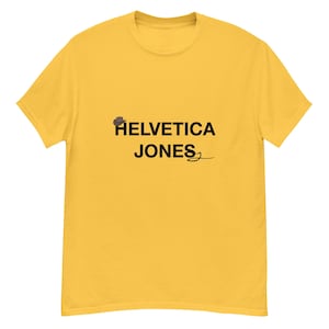 May include: A yellow t-shirt featuring the black text "HELVETICA JONES", with a small hat graphic above "HELVETICA" and a stylized signature below "JONES". The shirt is made of a soft material.