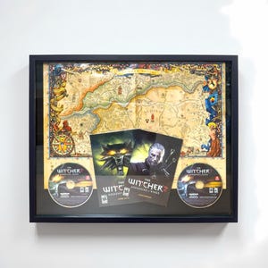 May include: Framed The Witcher 2 video game memorabilia. The display includes a map, game discs, and manuals. The map features a detailed, colorful design. The game discs and manuals are visible below the map, all encased in a black frame.