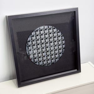 May include: Framed artwork featuring a circular microchip design. The chip is set against a black background within a black frame. The microchip has a grid-like pattern of small, rectangular components in shades of gray and white.