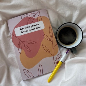 Peut inclure: Un planificateur rose et beige avec le texte "Ramadan planner & deen motivation" sur la couverture. Le planificateur est posé sur un couvre-lit blanc avec un stylo jaune et une tasse de café.