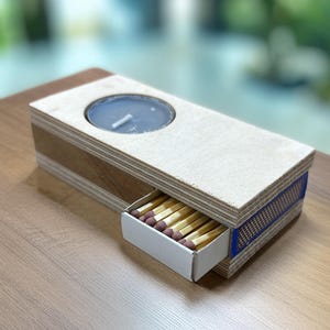 May include: A rectangular light-colored plywood candle holder featuring a circular cutout for a tealight candle. A small white drawer is partially pulled out from the side, revealing a collection of wooden matches with brown tips.