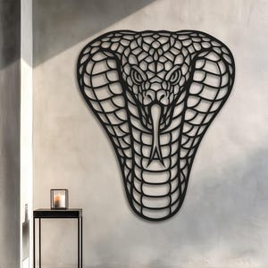 Può includere: Arte da parete a forma di cobra in metallo nero. Il cobra è raffigurato con intricati dettagli ritagliati, che mostrano la testa e il cappuccio del serpente. L'opera d'arte è montata su una parete di colore chiaro, creando un contrasto sorprendente.