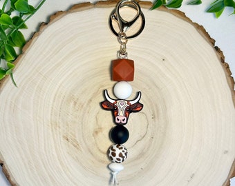 May include: A brown, white, and black cow charm keychain with a gold key ring. The charm features a cow head with a pink nose and white heart on its forehead. The keychain also includes a brown, white, and black patterned bead, a black bead, and a white bead.