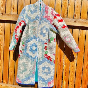 May include: A long-sleeved coat constructed from a quilted fabric featuring a patchwork pattern of hexagonal floral motifs in shades of light blue, pink, red, and white. The garment has a straight open front, a high collar, and a solid teal lining.