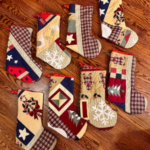 Puede incluir: Una colección de ocho calcetines navideños con diseños de patchwork. Cada calcetín presenta una combinación única de patrones, incluyendo cuadros, estrellas y aplicaciones temáticas navideñas. La paleta de colores incluye rojo, azul, beige y verde.