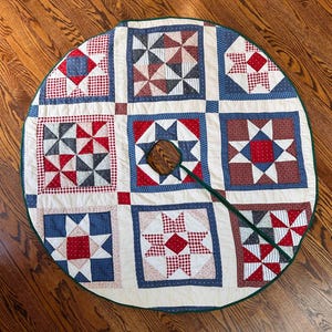 May include: A circular fabric tree skirt featuring a patchwork design of red, white, and blue geometric star and pinwheel patterns. The quilt-style textile has a dark green border and a central opening with a dark brown wooden fastener.