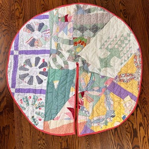 May include: A circular quilted tree skirt featuring a patchwork design of various floral and geometric fabric patterns in shades of purple, green, yellow, and white. The edge is finished with a thin red and white polka dot fabric trim.