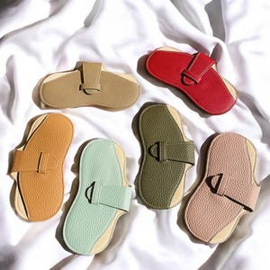 May include: A selection of leather eyeglass cases in shades of tan, red, orange, green, and pink. Each case has a contrasting interior and a strap closure with a metal buckle. The cases are displayed on a white fabric background.