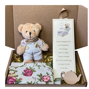 May include: A plush teddy bear in dungarees and a floral top is presented in a cardboard box with a floral fabric. A bookmark with a bear illustration and text is also included. The text on the bookmark reads "There's always time..."