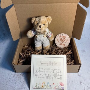 May include: A small, beige teddy bear wearing overalls and a floral shirt is in a brown cardboard box with shredded paper. A wooden tag with the text "A little Hug" is also in the box. A card with the text "Sending lots of love! I hear you've been poorly But now you're on the mend. So I'm sending you a little bear To be your Healing Friend!" is in the box.