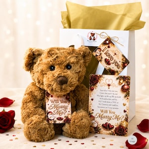 May include: A brown plush teddy bear with a tag reading Will You Marry Me, positioned in front of a white gift bag with gold tissue paper. A rectangular card features the text Will You Marry Me and a poem, alongside a tag reading I love you.