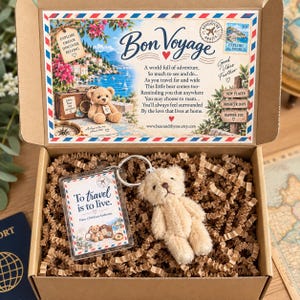 May include: A cardboard gift box containing a small beige plush teddy bear and a rectangular acrylic keyring. The keyring features the text 'To travel is to live.' The box lid interior displays a 'Bon Voyage' graphic with a poem and travel imagery.
