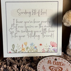 May include: A white card with a handwritten message that reads "Sending Lots of Love! I hear you've been poorly. But now you're on the mend. So I'm sending you a little bear to be your Healing Friend!"  The card is decorated with a floral border. A small wooden circle with the text "A little Hug" is also in the image.