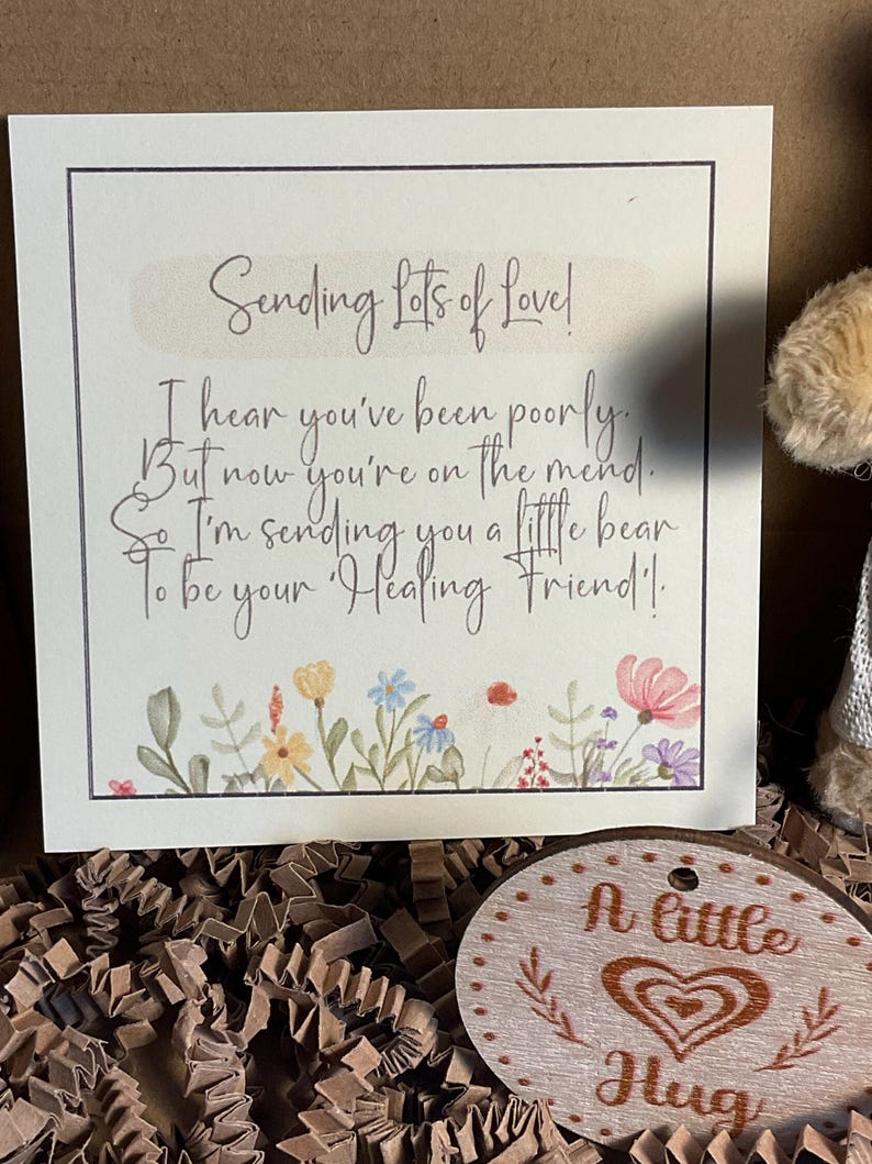 May include: A white card with a handwritten message that reads "Sending Lots of Love! I hear you've been poorly. But now you're on the mend. So I'm sending you a little bear to be your Healing Friend!"  The card is decorated with a floral border. A small wooden circle with the text "A little Hug" is also in the image.