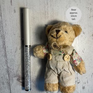 May include: A small, beige teddy bear wearing a white overall with a floral print shirt. The bear is approximately 10 inches tall. A silver glitter pen is in the background.