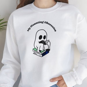 Puede incluir: Sudadera blanca con el texto "My Haunting Obsession" sobre un fantasma de dibujos animados leyendo un libro. El fantasma está sentado sobre una pila de libros con cubiertas verdes y azules. La sudadera tiene mangas largas.
